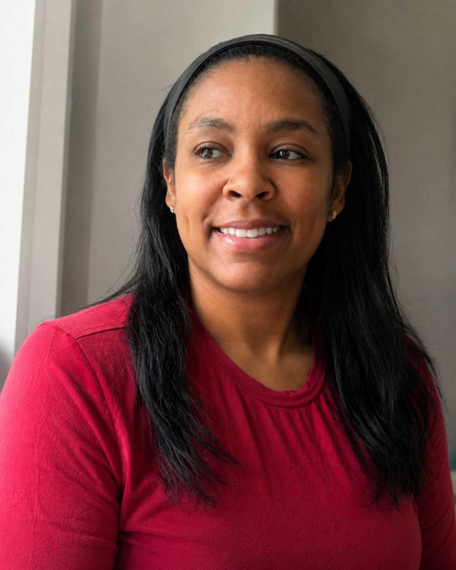 Jasmine Jones sits indoors near a window, wearing a bright red long-sleeve shirt, a black headband, and small stud earrings. She smiles softly while looking off to the side. Natural light from the window illuminates her face, with a neutral-colored wall in the background.