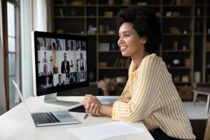 A woman sits at her desk and takes notes as she smiles at a group video chat